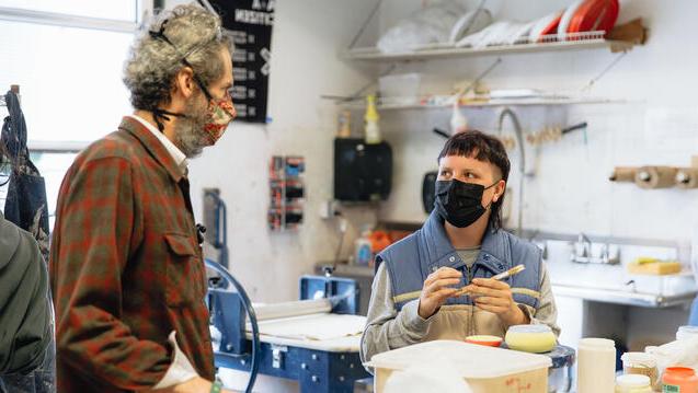 Instructor addresses student sitting at table with hand made ceramic item holding a paintbrush